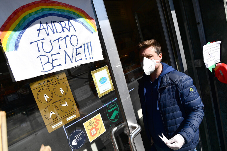 Una persona con la mascherina protettiva vicino ad un cartello  'Andra ' tutto bene ' - RIPRODUZIONE RISERVATA